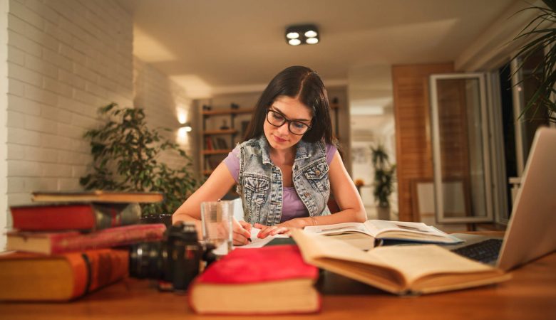 como focar nos estudos: mulher jovem sentada em mesa de casa em volta de vários livros