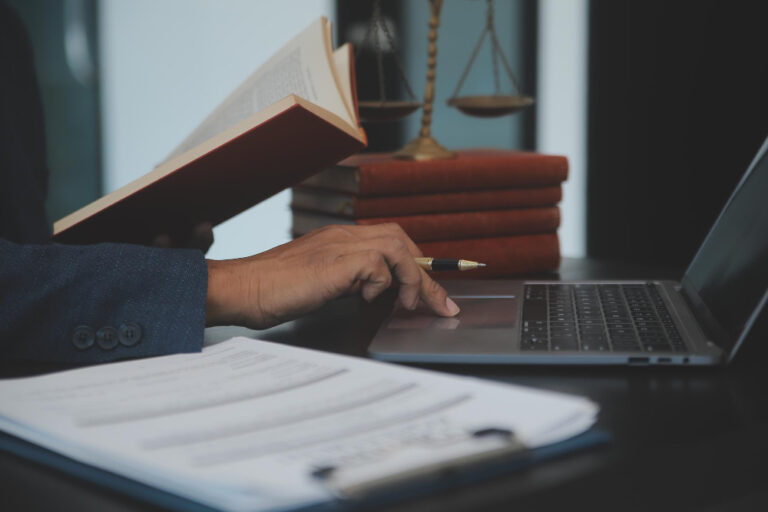 Mão operando um notebook com papéis, um livro e uma balança ao fundo